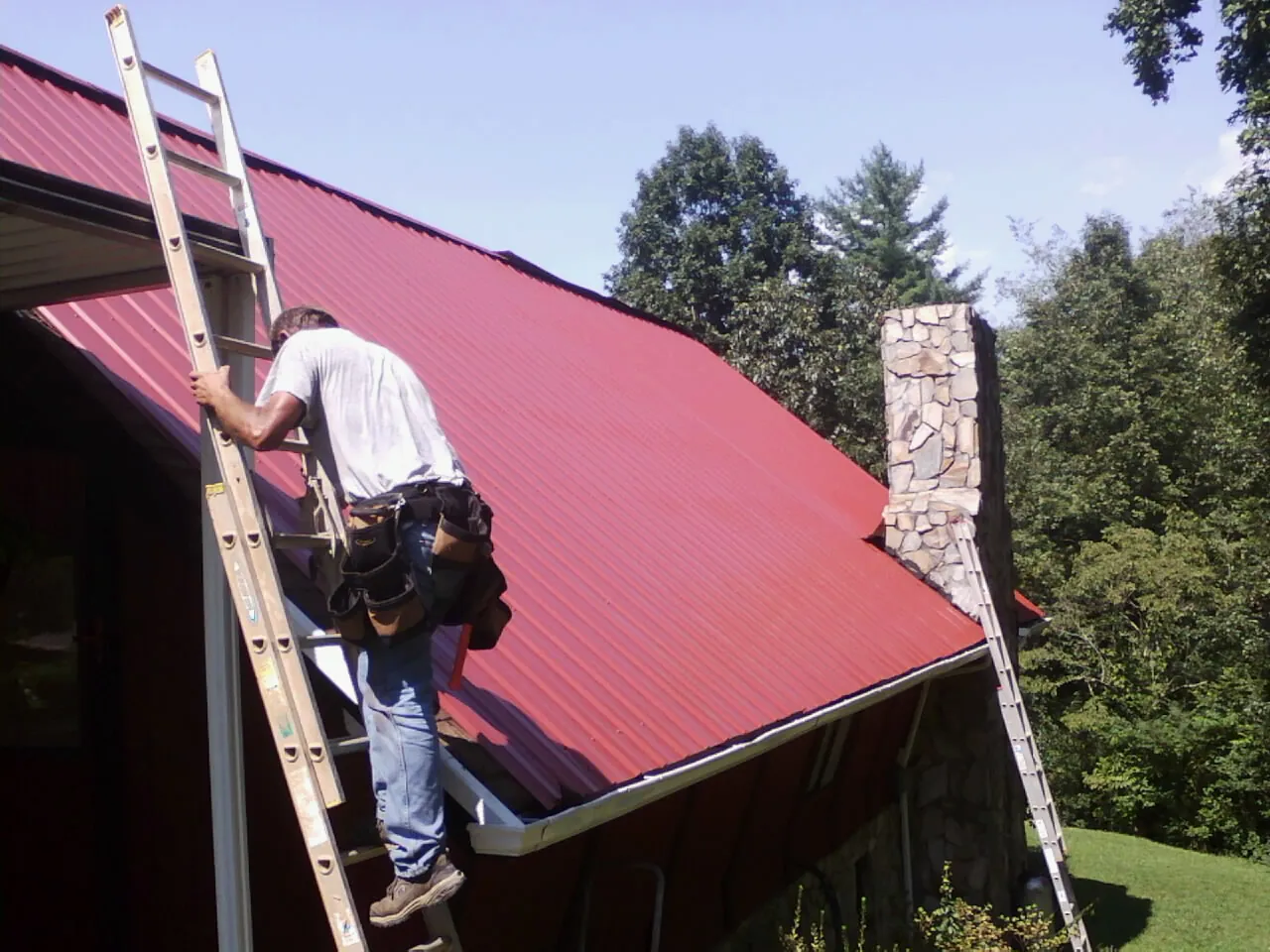 Roofer on a ladder beside a red metal roof and stone chimney — Soffit & Fascia Repair by Roofing Brothers in Mashantucket